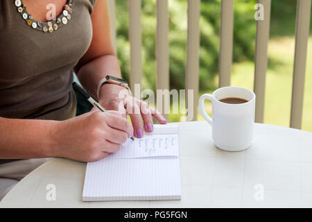 Una donna di mezza età a scrivere una lista della spesa su un tavolo esterno mentre gustate una tazza di caffè Foto Stock