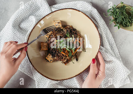 Vista parziale di mani femminili e soba con tofu e verdure decorate con semi germinati di girasole sul piano portapaziente grigio Foto Stock