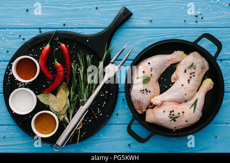 Vista dall'alto di cosce di pollo nella padella e spezie differenti sul tagliere con carne forcella sul piano portapaziente blu Foto Stock