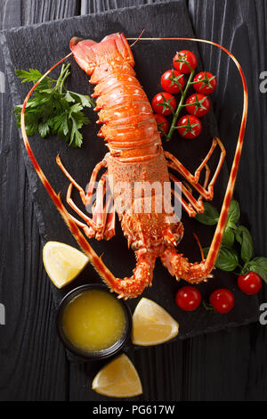 Lussuoso bollito di aragosta circondato da pomodori freschi, limone, erbe aromatiche e burro fuso close-up su una pietra nera. Verticale in alto vista da sopra Foto Stock