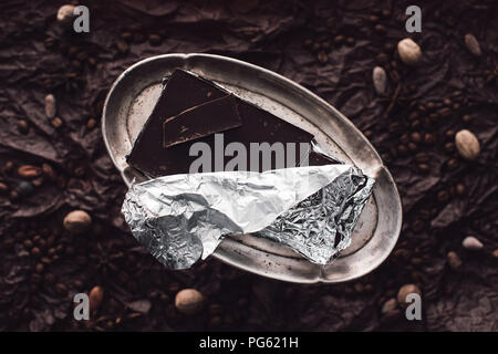 Vista in elevazione del cioccolato in lamina d'argento sul sentiero circondato da caffè in chicchi, le fave di cacao e noce moscata sulla superficie di copertura da black carta sgualcita Foto Stock