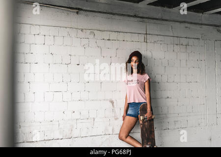 Ritratto di una giovane donna con lo skateboard, appoggiata contro la parete Foto Stock