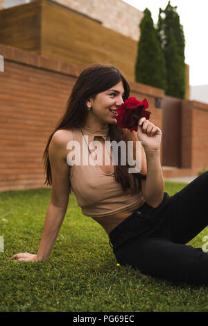 Sorridente giovane donna seduta su un prato odore di rose rosse Foto Stock