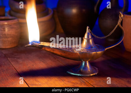 Bella antica olio lampada in bronzo arde su un tavolo di legno. Ceramica in background. Concetto di meraviglioso Aladdin's Lamp pieno di forze magiche Foto Stock