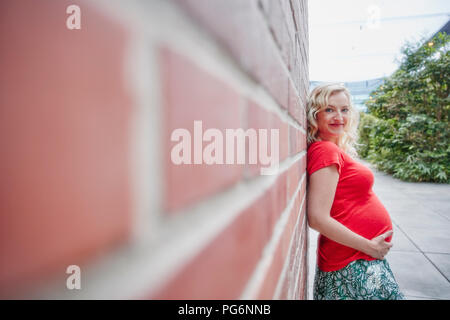 Ritratto di sorridere donna incinta all'aperto appoggiata contro un muro di mattoni Foto Stock