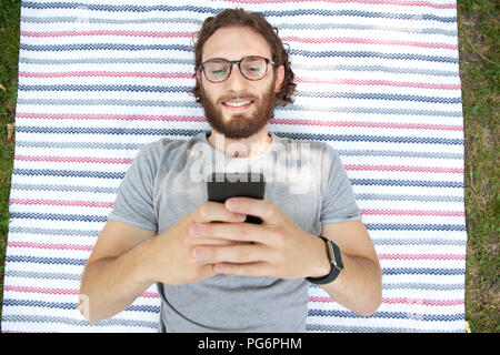 Ritratto di uomo sorridente giacente su una coperta in un parco tramite telefono cellulare, vista dall'alto Foto Stock