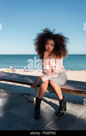 Ritratto di giovane e bella donna con taglio di capelli afro seduta su una panchina in spiaggia Foto Stock