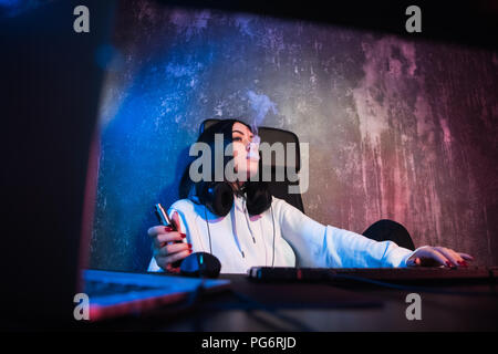 Giovane Hip brunette donna che indossa gli occhiali e la felpa con cappuccio, seduti da scrivania con computer mentre il fumo e-sigaretta Foto Stock