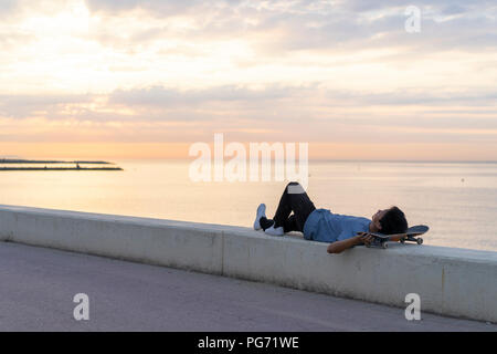 Giovane uomo cinese con lo skateboard giacente sulla parete in spiaggia Foto Stock