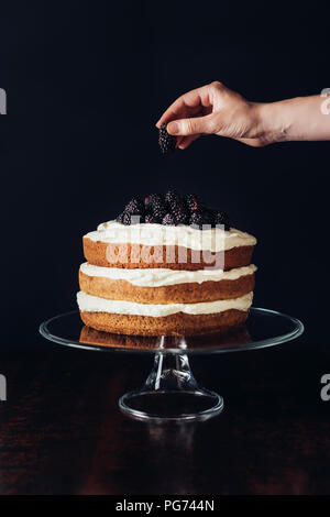 Ritagliato colpo di donna decorazione di gustosa torta blackberry sul piedistallo di vetro su nero Foto Stock