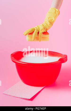 Immagine ritagliata femminile di detergente per il lavaggio di contenimento spugna sulla bacinella di plastica con schiuma, sfondo rosa Foto Stock