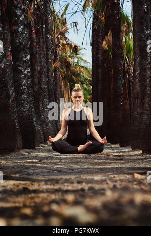 Giovane donna in nero sportswear seduti nella posizione del loto e meditando all'esterno. Donna in gestazione precoce. Pre natal esercitando, uno stile di vita sano. Foto Stock