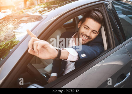 Ritratto di imprenditore guida in auto e sorridente sul suo tragitto per recarsi al lavoro. Giovane uomo il suo dito puntato verso la fotocamera. Foto Stock