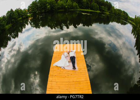 Coppia di sposi si posa su un molo in legno sullo sfondo del lago di superficie con riflettere di cielo nuvoloso Foto Stock