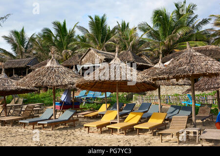 Sedie a sdraio sotto gli ombrelloni di paglia sulla sabbia in una spiaggia Bang, nel Vietnam centrale. La città costiera è situato vicino all'UNESCO protetti città di Hoi Foto Stock