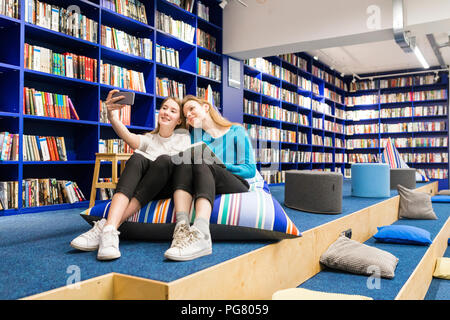 Due ragazze adolescenti seduti su beanbag in una biblioteca pubblica tenendo selfie con lo smartphone Foto Stock