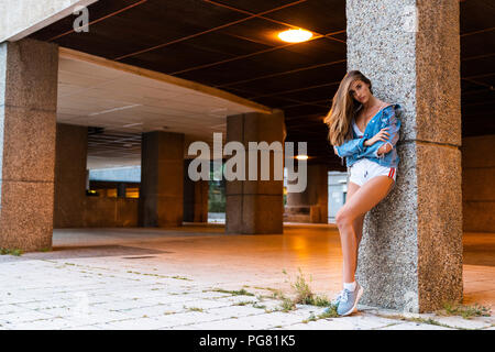 Attraente giovane donna in abbigliamento sportivo e giacca denim appoggiata contro una colonna Foto Stock