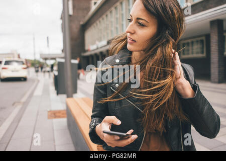 Giovane donna con capelli spazzate dal vento tenendo il cellulare in città Foto Stock