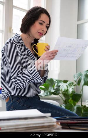 Donna con la tazza di caffè guardando carta alla scrivania in ufficio Foto Stock