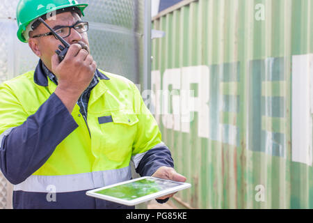 Lavoratore con tablet indossando giubbotto di sicurezza utilizzando walkie talkie in corrispondenza del recipiente Foto Stock