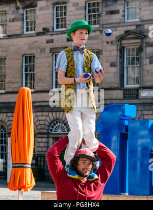 Edinburgh Fringe Festival di Edimburgo, Scozia, Regno Unito. 24 agosto 2018. Il sole splende artisti di strada presso la Vergine denaro sponsorizzato street sede sul Royal Mile. Un ragazzo giovane vestito in un costume colorato in piedi su un uomo di spalle come si destreggia sfere Foto Stock