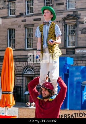 Edinburgh Fringe Festival di Edimburgo, Scozia, Regno Unito. 24 agosto 2018. Il sole splende artisti di strada presso la Vergine denaro sponsorizzato street sede sul Royal Mile. Un ragazzo giovane vestito in un costume colorato in piedi su un uomo di spalle come si destreggia sfere Foto Stock