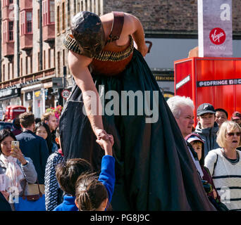 Edinburgh Fringe Festival di Edimburgo, Scozia, Regno Unito. 24 agosto 2018. Il sole splende sui frequentatori della frangia e musicisti del festival presso la Vergine denaro sponsorizzato street sede sul Royal Mile. Un uomo in un costume spaventoso che indossa un lungo mantello nero, casco, parte superiore in pelle e cinghia bullet passeggiate su palafitte tra la folla festival distribuendo volantini per il Fringe mostrano il silenzio e dà un giovane ragazzo in una stretta di mano Foto Stock