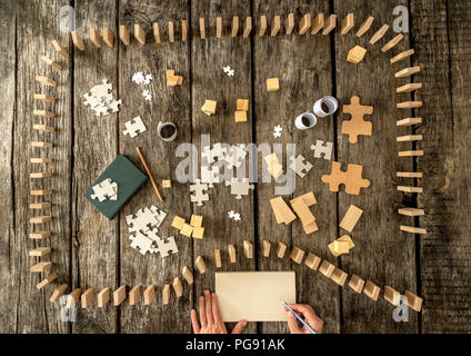 Vista dall'alto in basso di domino e i pezzi del puzzle su tavole di legno. Mano di scrittore con la penna e il blocco note. Foto Stock