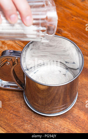La cottura di una torta - versare la farina dal bicchiere in acciaio spolverizzatore Foto Stock