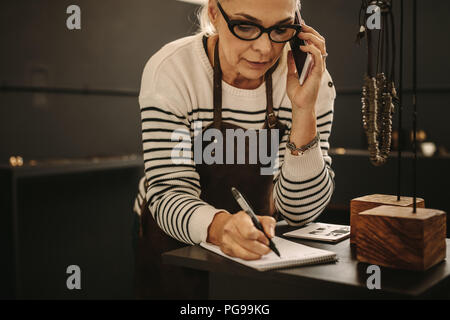 Ritratto di femmina matura gioielliere parlando al telefono cellulare e a rendere note. Goldsmith iscritto in un libro mentre si effettua una chiamata telefonica presso il suo studio di design Foto Stock