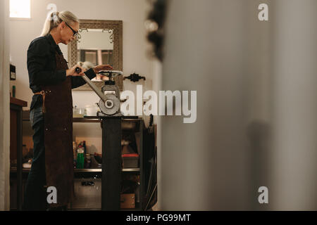Senior orafo femmina creazione di metallo sulla macchina di laminazione in officina. Donna matura lavorando nel laboratorio orafo. Foto Stock