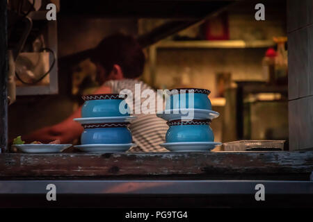Blu piatti di terracotta con cibi caldi in loro in attesa di essere prelevato dal cameriere su una finestra della cucina, il cuoco visibile sfumata in background Foto Stock