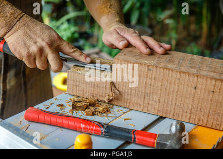Uomo in grembiule del cesello di legno con il bulino su una sega da tavolo. Posa di martello sul tavolo. Mani coperte in segatura. Attrezzi di falegnameria, concetto di lavorazione del legno. Foto Stock
