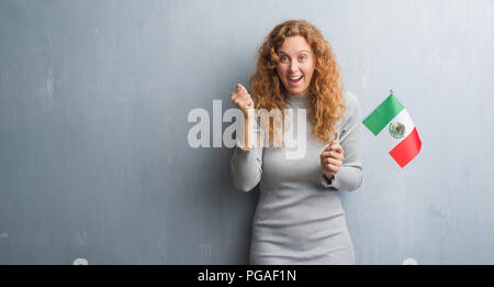 Giovane donna rosso su grigio parete grunge holding Bandiera del Messico urlando orgoglioso e celebrando la vittoria e il successo molto eccitato, allietarla emozione Foto Stock