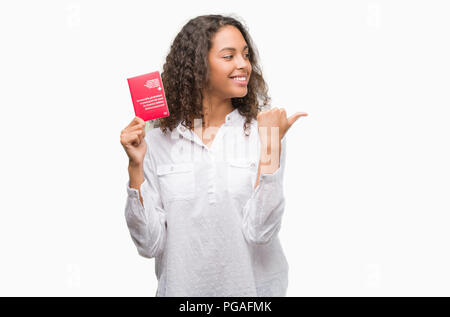 Giovane donna ispanica tenendo il passaporto della Svizzera e di puntamento che mostra con il pollice fino a che il lato con buon viso sorridente Foto Stock