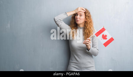 Giovane donna rosso su grigio parete grunge holding bandiera del Canada ha sottolineato con la mano sulla testa, sconvolto con vergogna e la sorpresa di fronte, arrabbiato e vanificare Foto Stock