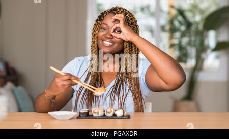 African American donna mangiare sushi con buon viso sorridente facendo segno OK con la mano sull'occhio guardando attraverso le dita Foto Stock