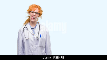 Giovani redhead donna che indossa uniformi medico con la lingua fuori con felice espressione divertente. Concetto di emozione. Foto Stock