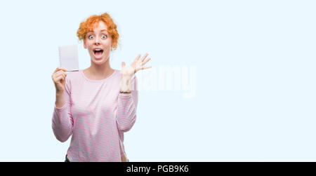 Giovane donna redhead holding notebook bianco molto felici ed eccitati, vincitore espressione celebrando la vittoria urlando con un grande sorriso e le mani alzate Foto Stock