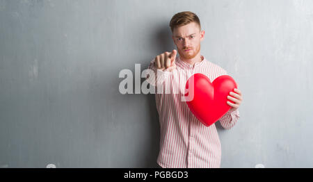 Giovani redhead man over grigio parete grunge holding cuore rosso puntando con il dito per la telecamera e a voi, mano segno, positivo e fiducioso gesto fro Foto Stock