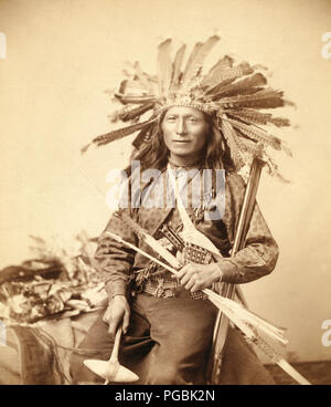 La foto mostra il piccolo, Oglala band leader, tre quarti di lunghezza ritratto in studio, seduto, indossando una Turchia di acconciatura di piume e tenendo premuto varie armi. Foto Stock