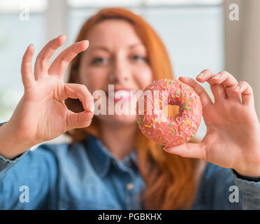 Redhead donna ciambella di contenimento a casa facendo segno ok con le dita, simbolo eccellente Foto Stock