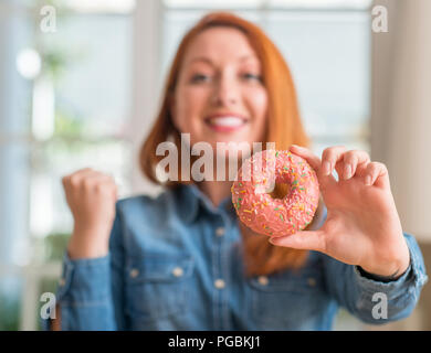 Redhead donna ciambella di contenimento a casa urlando orgoglioso e celebrando la vittoria e il successo molto eccitato, allietarla emozione Foto Stock