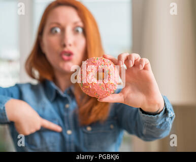 Redhead donna ciambella di contenimento a casa con sorpresa faccia puntare il dito a se stesso Foto Stock