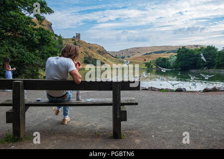 Edinburgh, Regno Unito - 27 Luglio 2018: il giovane uomo si siede e fuma su un banco da St Margaret Loch con le rovine della basilica di Sant'Antonio cappella del backgro Foto Stock