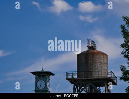 Molto vecchio parzialmente arrugginito water tower con un clock tower in primo piano e cielo blu in background Foto Stock