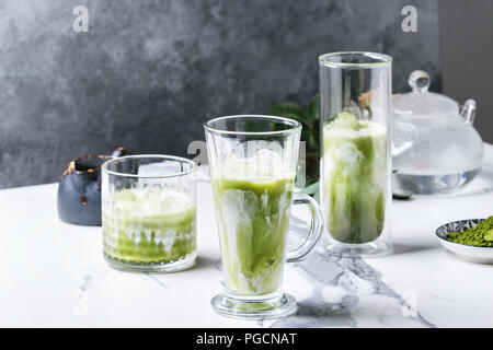Tè verde Matcha iced latte macchiato o un cocktail in tre diversi tipi di bicchieri con cubetti di ghiaccio, matcha polvere e il contenitore del latte su bianco tavola di marmo, decorata da g Foto Stock