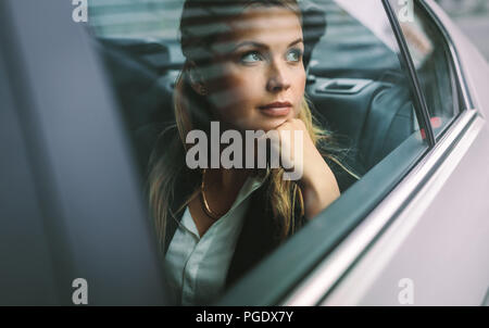 Bella giovane imprenditrice seduta sul sedile posteriore di una macchina e guardando fuori dalla finestra. Femmina executive aziendali che viaggiano da una cabina. Foto Stock