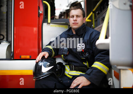 Foto del giovane vigile del fuoco con il casco in tuta siede in camion dei pompieri Foto Stock