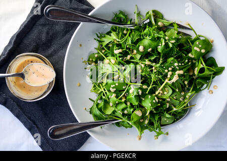 Flat-lay insalata verde di mizuna e minatore della lattuga servita con il rabarbaro e semi di papavero condimento, servita in una ciotola di bianco con due cucchiai di portata. Foto Stock